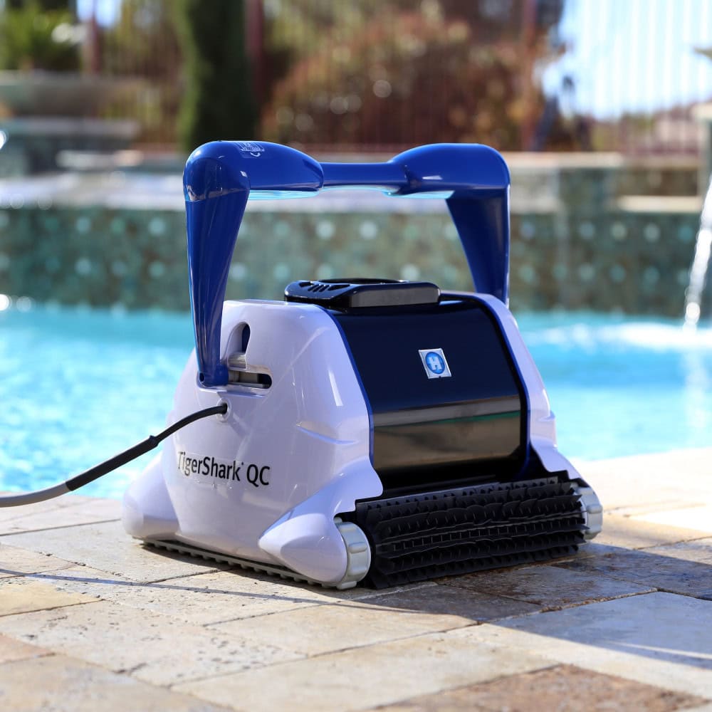 A TigerShark QC robotic pool cleaner rests at the sunlit edge of a clean swimming pool.