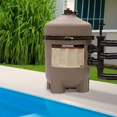 A Hayward pool filter sits next to a clean swimming pool, surrounded by green plants and a nearby building.