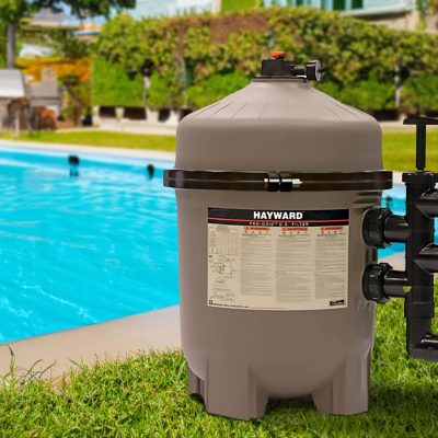 A large Hayward pool filter stands on grass beside a bright blue backyard swimming pool in sunlight.