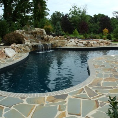 A solid black swimming pool with a stone patio, waterfall, lounge chairs, and trees in Long Island.