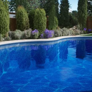 A bright blue pool with New Hampton Bays Tile/Sierra Floor, lush greenery, and purple flowers nearby.