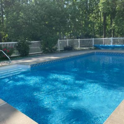 A girl in blue sits on Luxe Gemstone Tile Ocean Breeze floor by a pool, trees, and white fence in Queens.