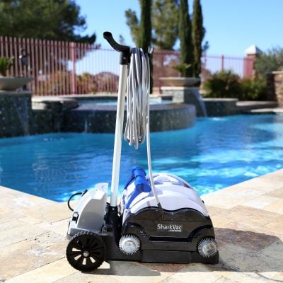 A SharkVac pool cleaner with hose and handle rests on poolside tiles beside a clear blue swimming pool.