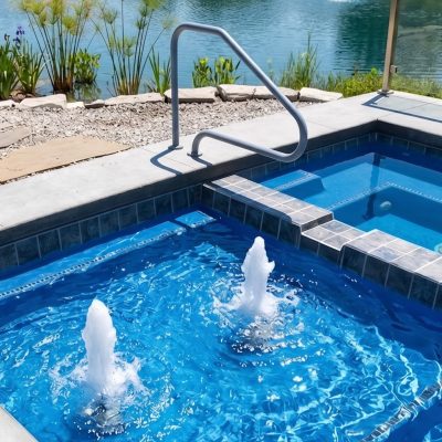 A small pool with a TruFit Bubbler, steps and metal railing, sits near a lake with lush plants behind.