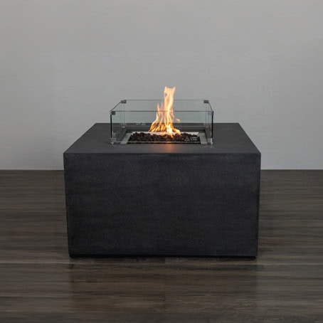 The 42″ Square Liquid Propane Fire Bowl with glass wind guard sits on dark wood flooring by a plain wall.